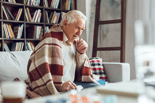 an older man sitting on a couch wrapped with a blanket and coughing